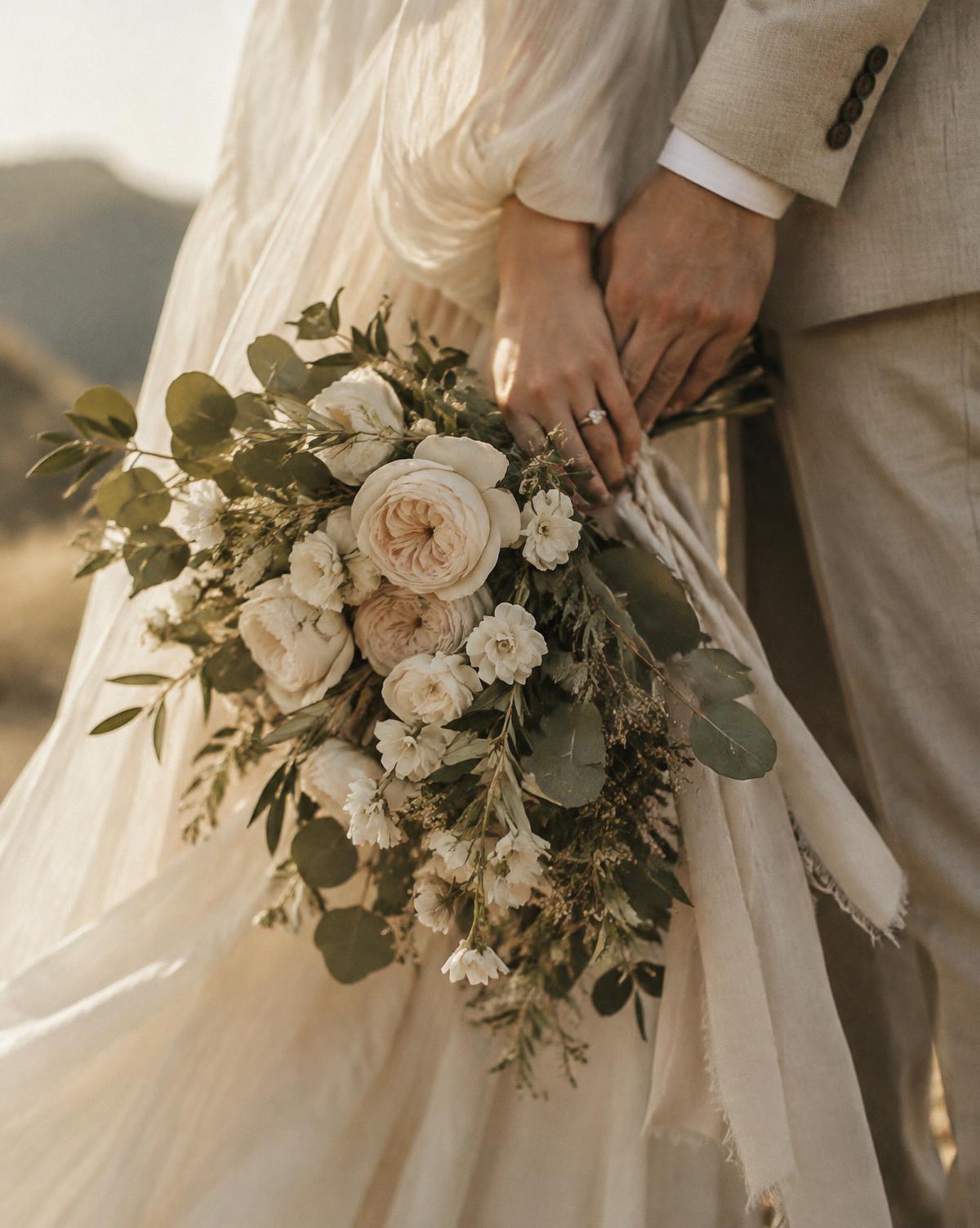 A couple holding hands beside a bouquet of ivory garden roses and eucalyptus